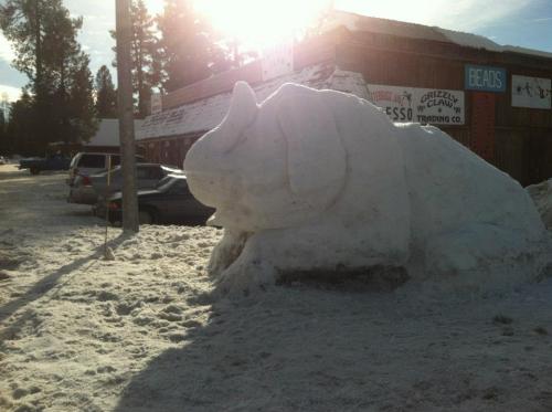 A large snow sculpture resembling a dog sits in a snowy area near a building, with trees and cars in the background.