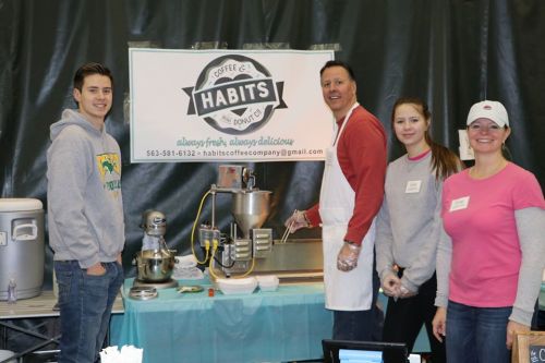A group of four people stands behind a table with a sign for "Habits Coffee & Donut," preparing food and drinks.