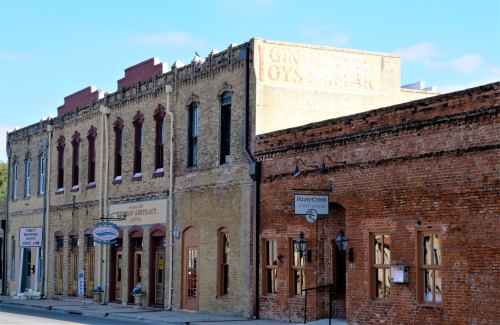 Historic brick buildings line a street, featuring signs for a bar and an antique shop under a clear blue sky.