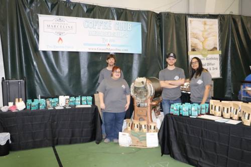 A group of four people at a coffee club booth, showcasing products and a coffee roaster in a market setting.
