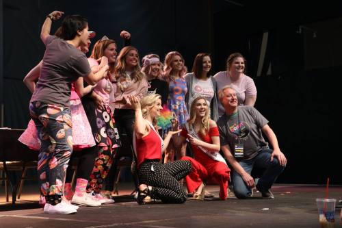 A group of people posing on stage, smiling and dressed in colorful outfits, with a mix of casual and formal attire.