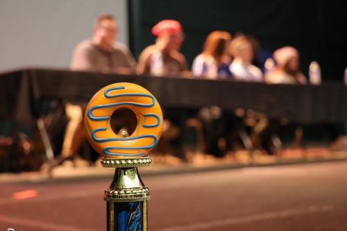 A close-up of a trophy with a donut design, with a blurred panel of judges in the background.