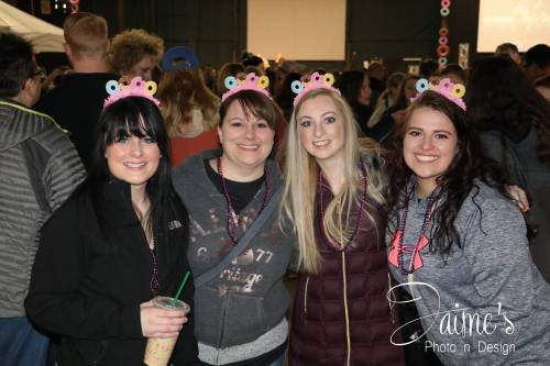Four women wearing colorful headbands smile together at a lively event, with a crowd and stage in the background.
