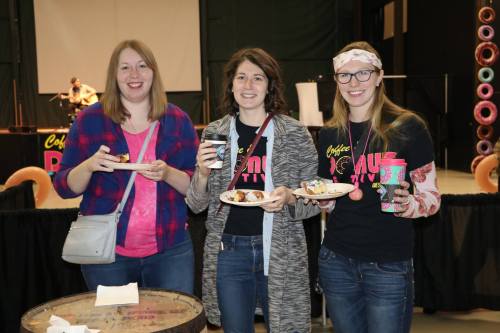 Three women hold plates of food and drinks, smiling at an event with a stage in the background.