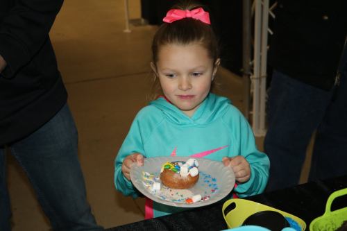 A young girl in a turquoise hoodie holds a decorated donut on a plate, looking thoughtfully at it.