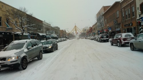 Snow-covered street lined with parked cars and festive decorations, creating a winter scene in a small town.