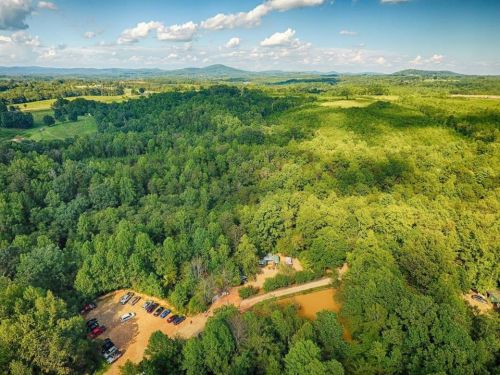 Aerial view of lush green forest with a parking area and distant mountains under a blue sky with scattered clouds.