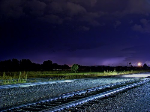 Nighttime scene of railway tracks under a dark sky, with distant lights and silhouettes of trees in the background.