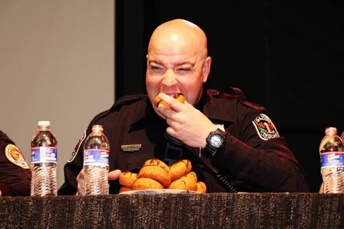 A police officer sitting at a table, eating a donut with bottles of water in front of him.