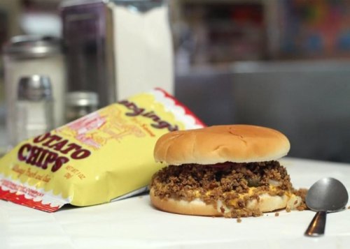 A sandwich filled with seasoned ground meat sits next to a bag of potato chips on a table.