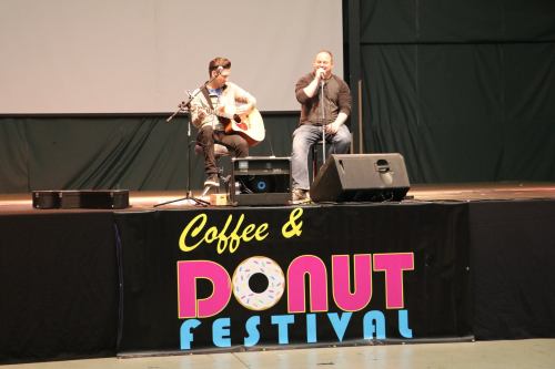 Two performers on stage at a Coffee & Donut Festival, one playing guitar and the other singing into a microphone.