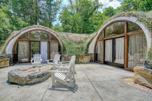 Two unique, grass-roofed structures with large windows, surrounded by trees and a stone patio with white chairs and a fire pit.