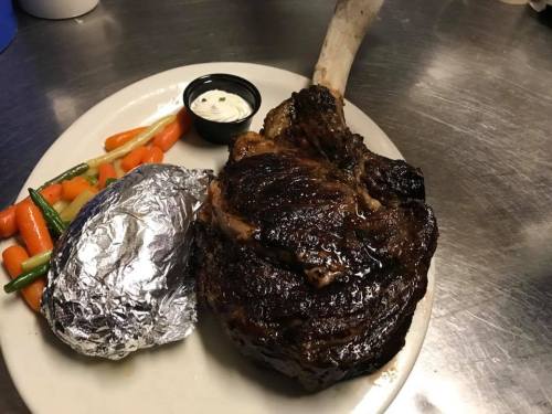 A large, juicy ribeye steak on a plate with a baked potato and mixed steamed vegetables on the side.