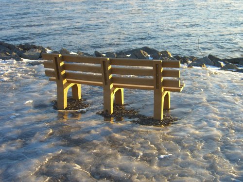 Two wooden benches partially submerged in icy water by the shore, with a calm sea in the background.