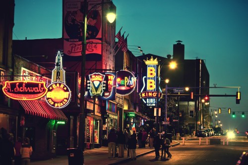 Colorful neon signs illuminate a lively street scene at dusk, featuring bars and music venues.