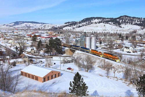 A train travels through a snowy landscape, passing a small town with hills in the background and silos nearby.