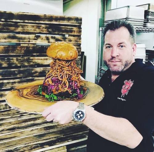 A chef holds a towering burger topped with crispy onion rings on a wooden platter, set against a rustic background.