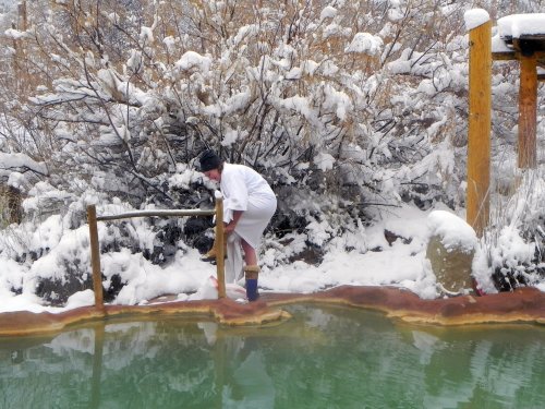 A person in white clothing stands by a hot spring surrounded by snow-covered trees and a winter landscape.