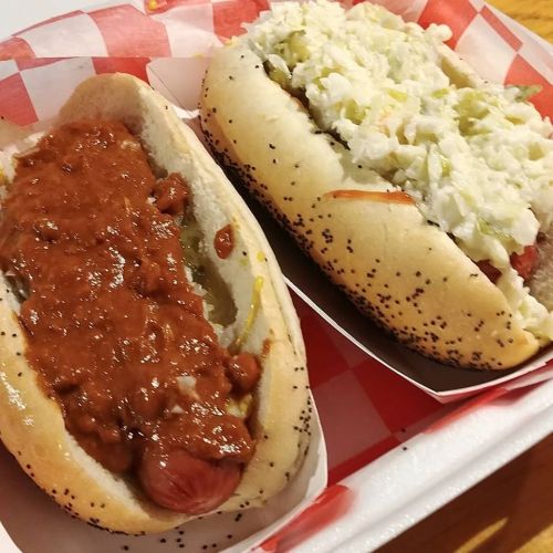 Two hot dogs in poppy seed buns: one topped with chili and the other with coleslaw, served in a red-checkered tray.