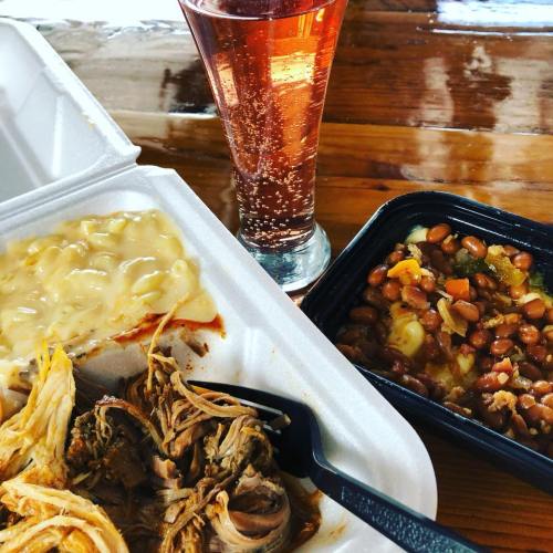 A takeout meal featuring pulled pork, macaroni and cheese, baked beans, and a glass of sparkling drink.