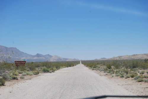 A dusty road stretches into the distance, flanked by sparse vegetation and mountains under a clear blue sky.