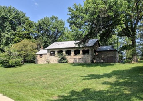 A stone house with a metal roof, surrounded by green grass and trees under a clear blue sky.