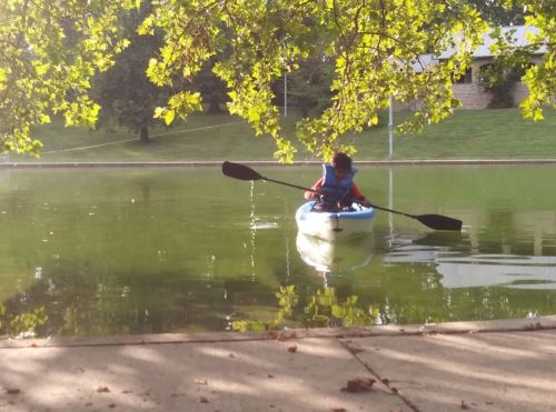 A person paddles a kayak on a calm lake, surrounded by green trees and reflecting water.