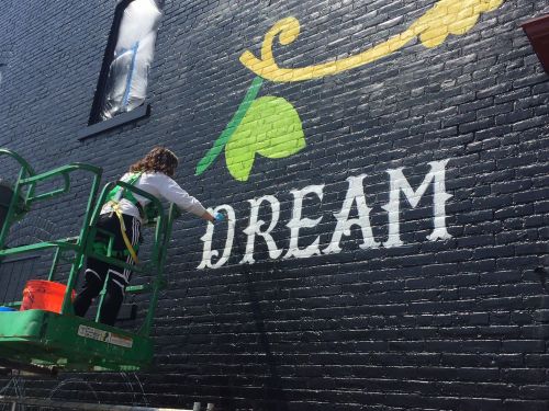 A person on a lift painting the word "DREAM" on a black brick wall, with green accents and a bright atmosphere.
