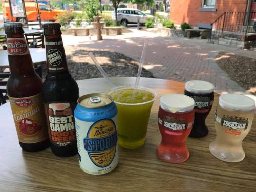A table with various drinks: two bottles of beer, a can of ale, a cup of yellow beverage, and colorful shot glasses.