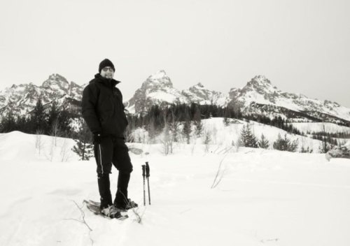 A person stands on snowshoes in a snowy landscape, with mountains and trees in the background. Black and white photo.