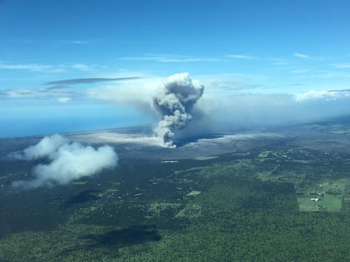 Aerial view of a volcano erupting, with a large plume of smoke rising above green landscape and ocean in the background.