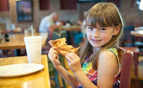 A smiling girl holds a slice of pizza in a restaurant, with a drink and empty plate on the table.