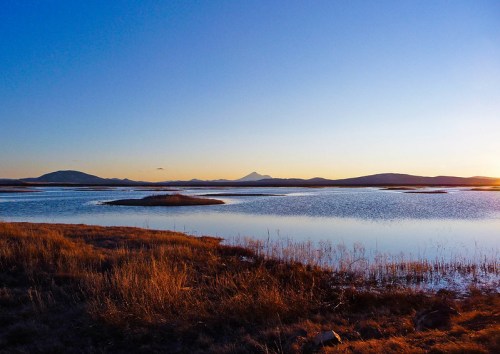 A serene landscape featuring a calm lake, distant mountains, and a clear blue sky at sunset.