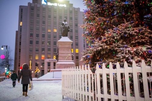 A snowy scene with a person walking past a decorated Christmas tree and a statue, with buildings in the background.