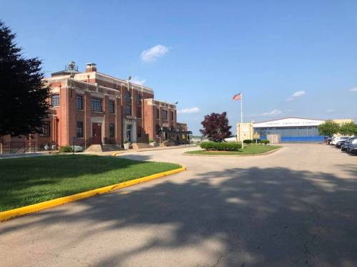 A brick building with an American flag, surrounded by greenery and a parking lot, under a clear blue sky.