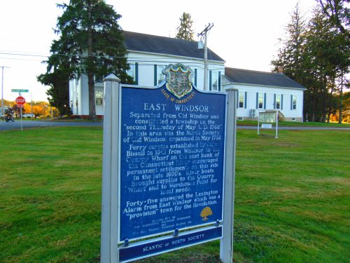 Historical marker for East Windsor, Connecticut, detailing its establishment and significance, with a church in the background.