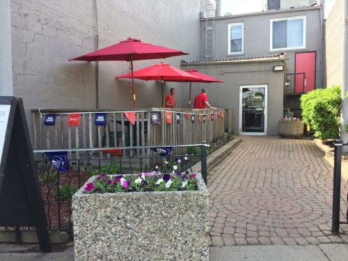 A patio with red umbrellas, two people in red shirts, and flower planters along a brick pathway.