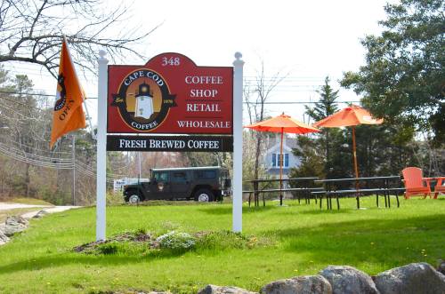 Sign for Cape Cod Coffee with flags, outdoor seating, and a jeep in the background on a sunny day.