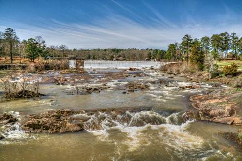 A serene river scene with flowing water, rocks, and a distant dam surrounded by trees under a clear blue sky.
