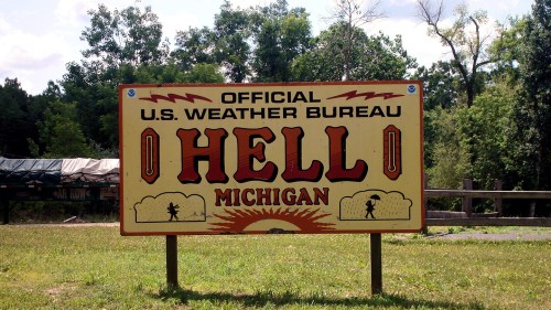 Sign reading "Official U.S. Weather Bureau Hell, Michigan" with a colorful design and trees in the background.