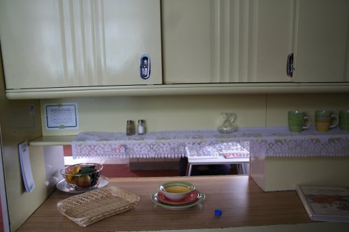 A vintage kitchen with yellow cabinets, a lace tablecloth, dishes, a fruit bowl, and a glass pitcher on a wooden countertop.