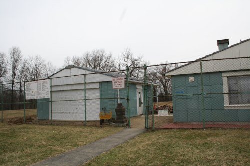 A fenced area with two small buildings, a sign, and a pathway, surrounded by bare trees and grass.