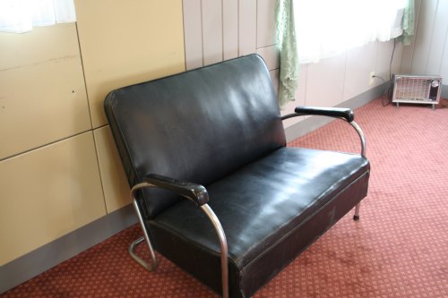 A black leather loveseat with metal arms, placed in a room with a patterned carpet and a window with sheer curtains.