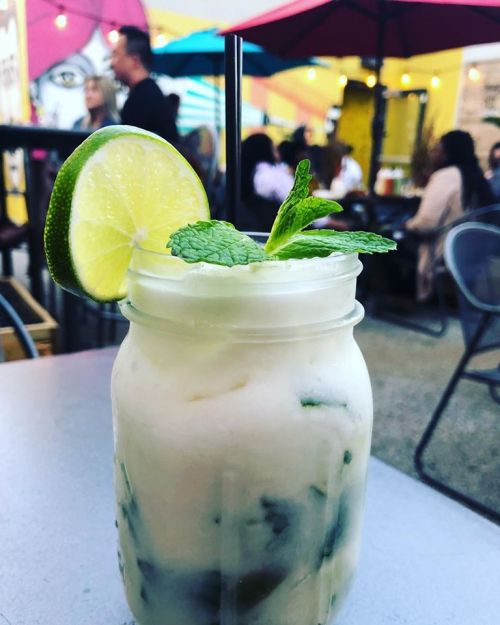 A refreshing drink in a jar with mint and lime, set on a table with people and colorful umbrellas in the background.