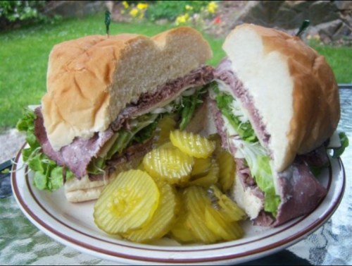 A large sandwich filled with roast beef and lettuce, served with a side of sliced pickles on a plate.