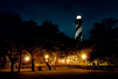 A lighthouse illuminated at night, surrounded by trees and soft glowing lights, creating a serene atmosphere.