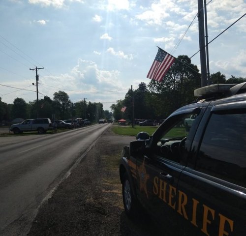 A sheriff's vehicle parked on a road lined with American flags and cars, under a bright sky with scattered clouds.