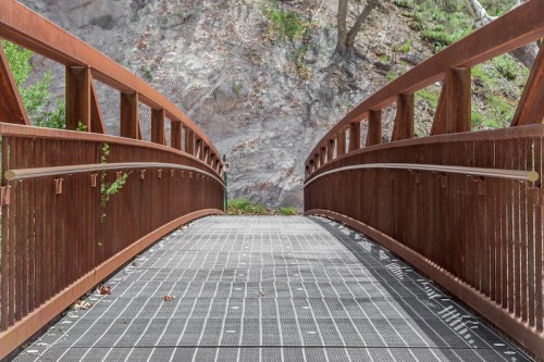 A rust-colored metal bridge spans a path, surrounded by greenery and a rocky hillside.