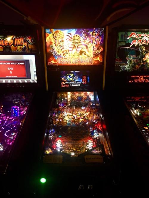 A dimly lit arcade featuring a pinball machine with an Iron Maiden theme and colorful lights.