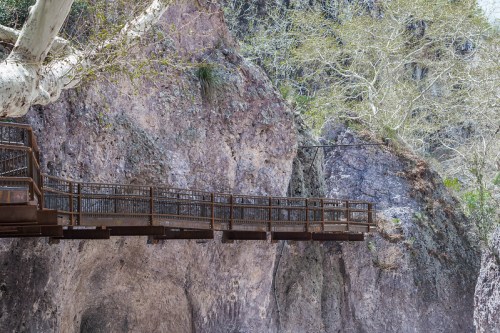 A wooden bridge spans a rocky landscape, surrounded by trees and steep cliffs.